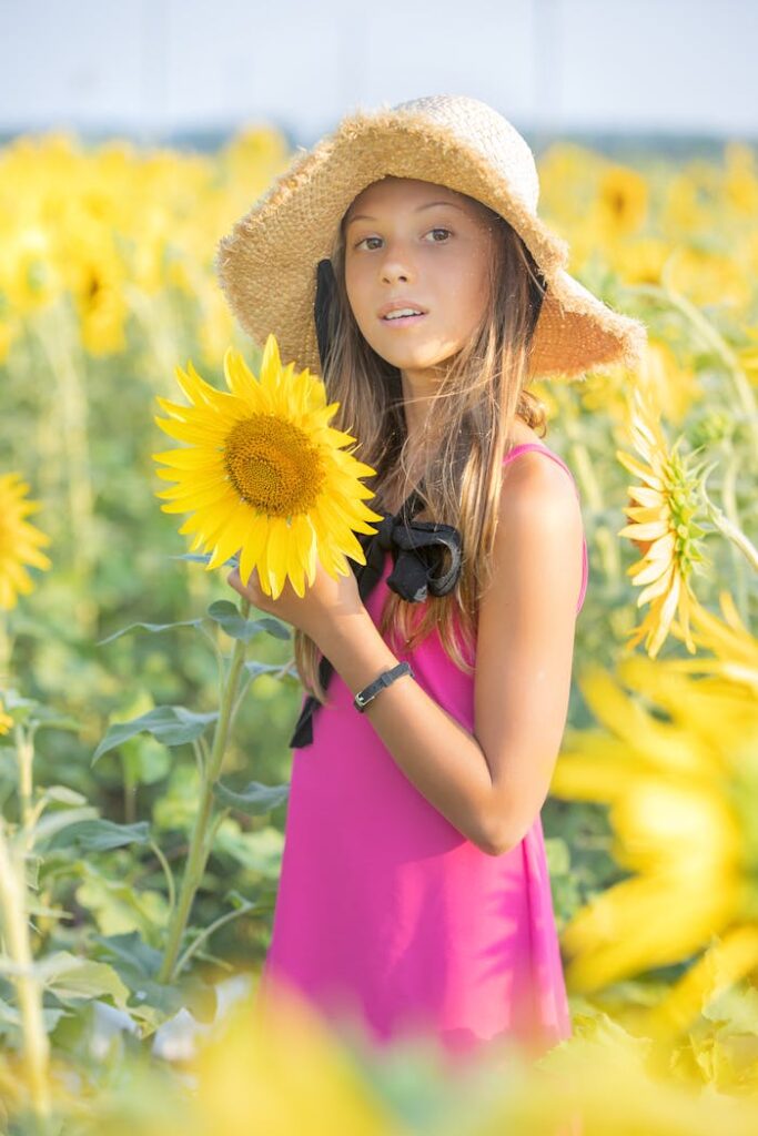A young girl in a pink dress holds a sunflower in a bright sunflower field during a sunny day.